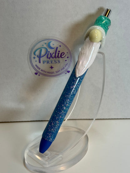 A Pixlie Press glitter pen featuring a Gnome figure with a blue glittery body, white beard,  displayed on a clear acrylic stand.