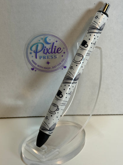 A Pixlie Press retractable pen with a white barrel decorated with black illustrations of open books, coffee cups, stars, and dots, resting on a clear acrylic stand.