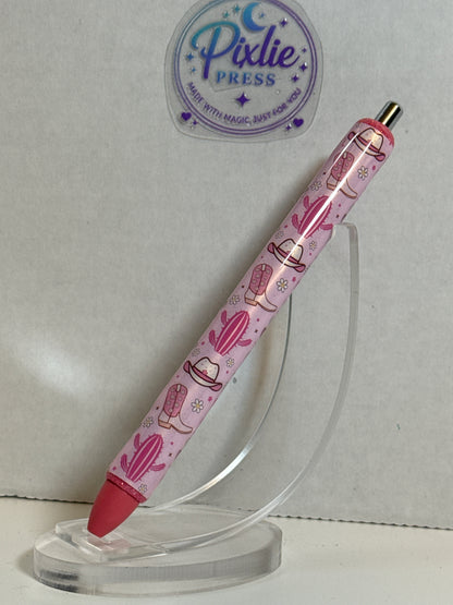 A pink Pixlie Press gel pen with a desert-themed print of cacti, hats, and a cowboy boot, displayed on a clear acrylic stand against a light gray background.