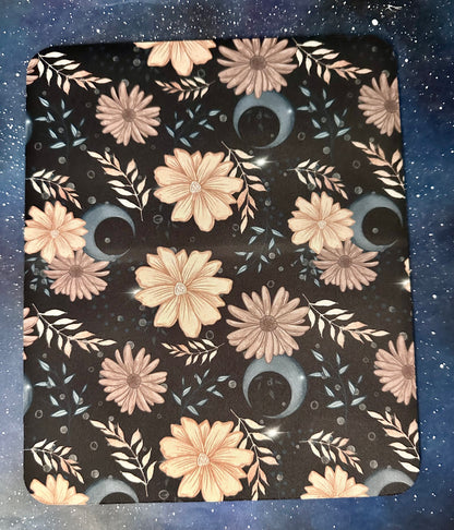 A square mouse pad with rounded corners featuring a celestial floral pattern on a black background, with pink and beige flowers, blue moon phases, and white leaves, shown on a blue starry space background.