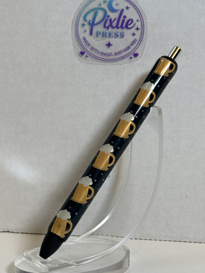 A black retractable pen with a repeating pattern of gold beer mugs with white foam, displayed on a clear acrylic stand against a light gray background with a Pixlie Press logo sticker.