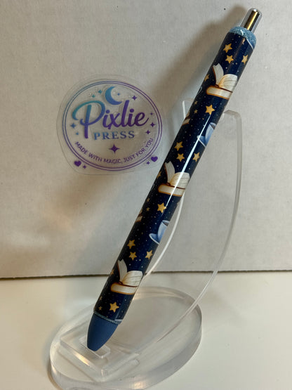 A Pixlie Press ballpoint pen with a dark blue space-themed design featuring stars, planets, and a rocket, displayed on a clear acrylic stand.